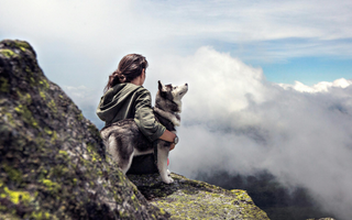 Woman sits on a mountaintop with her Siberian Husky, gazing at a cloudy vista.
