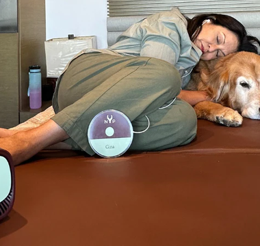 Woman relaxing on a brown mat with her dog, using a Gina device.  NYP logo visible.
