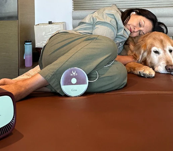 Woman relaxing on a brown mat with her dog, using a Gina device.  NYP logo visible.