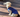 Adorable cream-colored longhaired dachshund puppy wearing a blue sweater sits on a wooden deck.