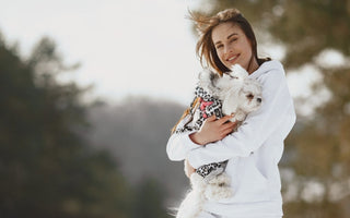 Happy woman in white hoodie cuddling small white dog in winter coat outdoors.