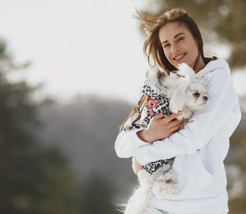 Happy woman in white hoodie cuddling small white dog in winter coat outdoors.