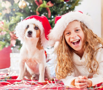 Happy girl and puppy in Santa hats by a Christmas tree, considering a puppy as a gift.
