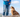 A person in jeans and a blue jacket stands on a rock overlooking a valley, with a brown English Bulldog sitting beside them.