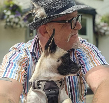 Man in hat and striped shirt cuddling small beige and black dog