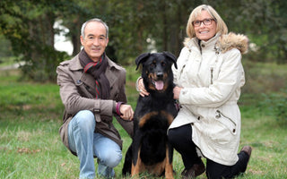 Happy senior couple kneeling with their Beauceron dog outdoors.
