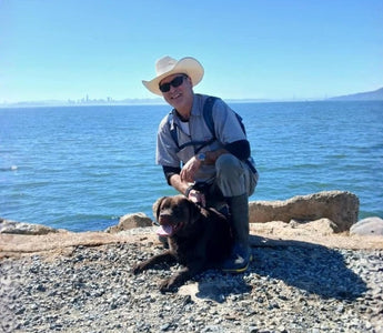 Man in cowboy hat with chocolate lab dog by the ocean, with a distant city skyline.