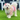 A small white fluffy dog wearing a pink and beige harness-style pet outfit with a ruffled pastel skirt, standing on green grass in front of white plastic chairs.